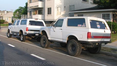 1996 Ford C-350 (Centurion Conversions) - Two white Ford trucks, a Bronco and an extended cab, are parked on a street. The Bronco has a custom bumper and off-road tires.