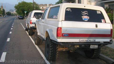 1996 Ford C-350 (Centurion Conversions) - Two white Ford Broncos are parked on the side of the road, one heavily modified with off-road features.
