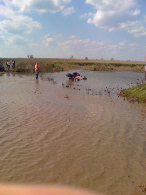 1990 Ford Bronco - Several people watch as an off-road vehicle is stuck in a muddy body of water under a cloudy sky.