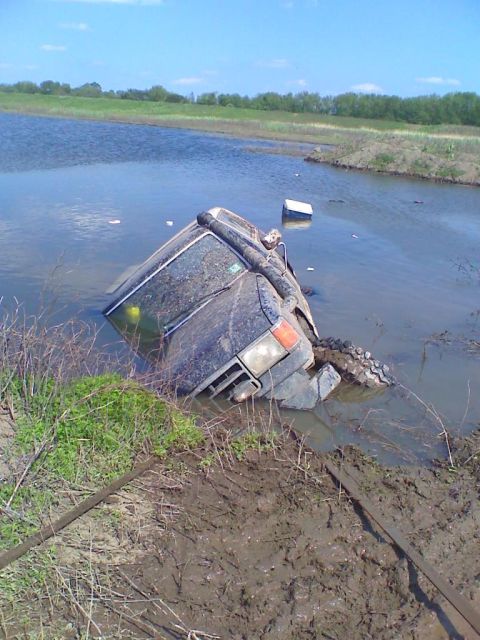 1990 Ford Bronco - A dark SUV is overturned and partially submerged in a body of water with muddy banks.