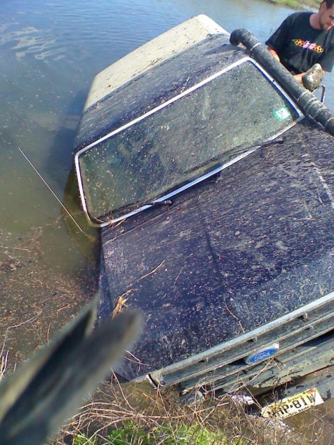 1990 Ford Bronco - A dark-colored Ford Bronco is stuck in murky water, with a person observing from the side.