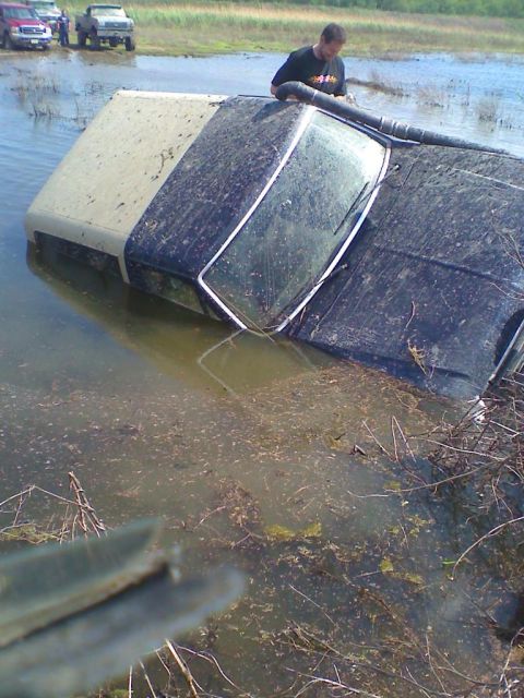 1990 Ford Bronco - A vehicle is overturned and partially submerged in murky water, with other vehicles visible in the background.