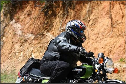 1985 Ford LTD Crown Victoria - A rider on a green Honda motorcycle navigates a road with a rocky hillside in the background.