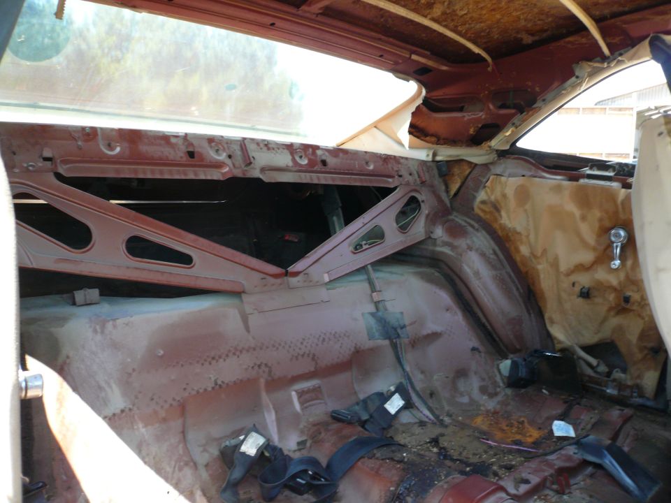 Interior view of a car with stripped-down panels, showing rust and exposed metal structure. Seatbelts are visible on the floor