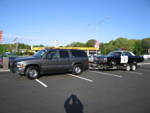 1991 Ford LTD Crown Victoria - gray Chevrolet Suburban tows a flatbed trailer carrying a black-and-white Highway Patrol sedan in a parking lot near a McDonald's.