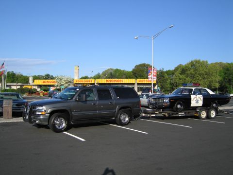 1991 Ford LTD Crown Victoria - gray Chevrolet Suburban with a trailer hauling a black-and-white police sedan, parked in a lot in front of a McDonald's.