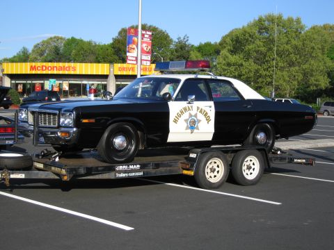 1991 Ford LTD Crown Victoria - black-and-white Highway Patrol sedan strapped onto a flatbed trailer in a parking lot in front of a McDonald's restaurant.