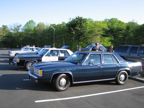 1991 Ford LTD Crown Victoria - dark-blue four-door sedan parked among a row of vintage marked police cars in a tree-lined parking lot.