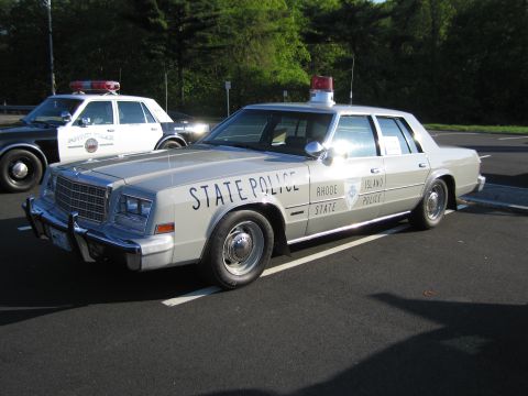 1991 Ford LTD Crown Victoria - silver Rhode Island State Police sedan parked in a lot beside a black-and-white patrol car under tall pines.