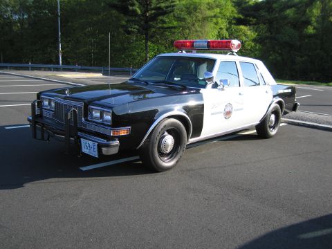 1991 Ford LTD Crown Victoria - Front three-quarter view of a black-and-white vintage police sedan with a red lightbar, parked alone in an empty lot.