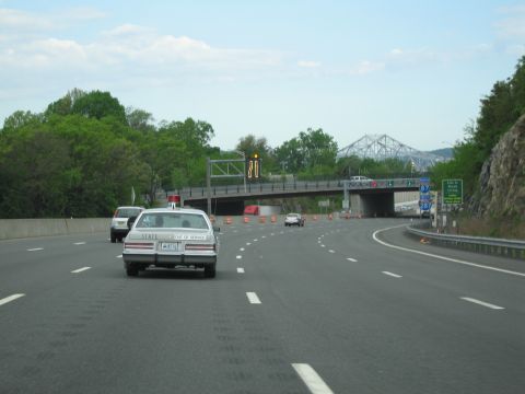 1991 Ford LTD Crown Victoria - white sedan with a State Police door decal drives along a multi-lane highway toward an overpass and a distant truss bridge.