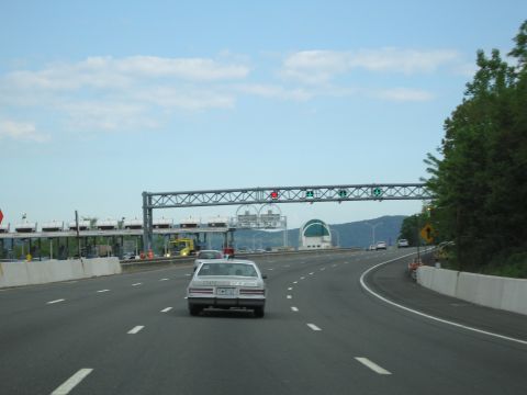 1991 Ford LTD Crown Victoria - white sedan travels along a highway approaching an overhead toll-plaza gantry with lane signals and toll booths ahead.