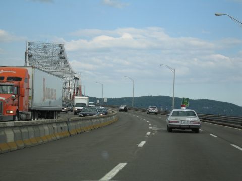 1991 Ford LTD Crown Victoria - white sedan drives across a bridge highway alongside an orange tractor-trailer with a steel truss span in the background.