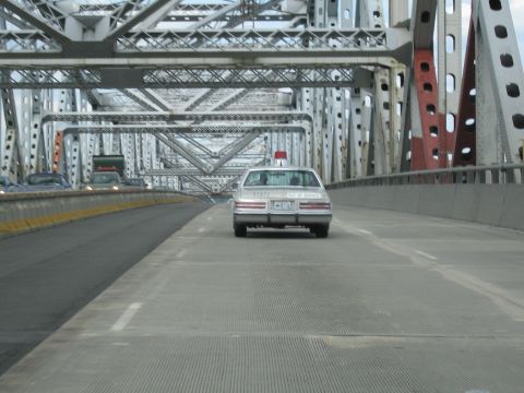 1991 Ford LTD Crown Victoria - silver police cruiser topped with a single red beacon travels across a steel truss bridge, viewed from behind.