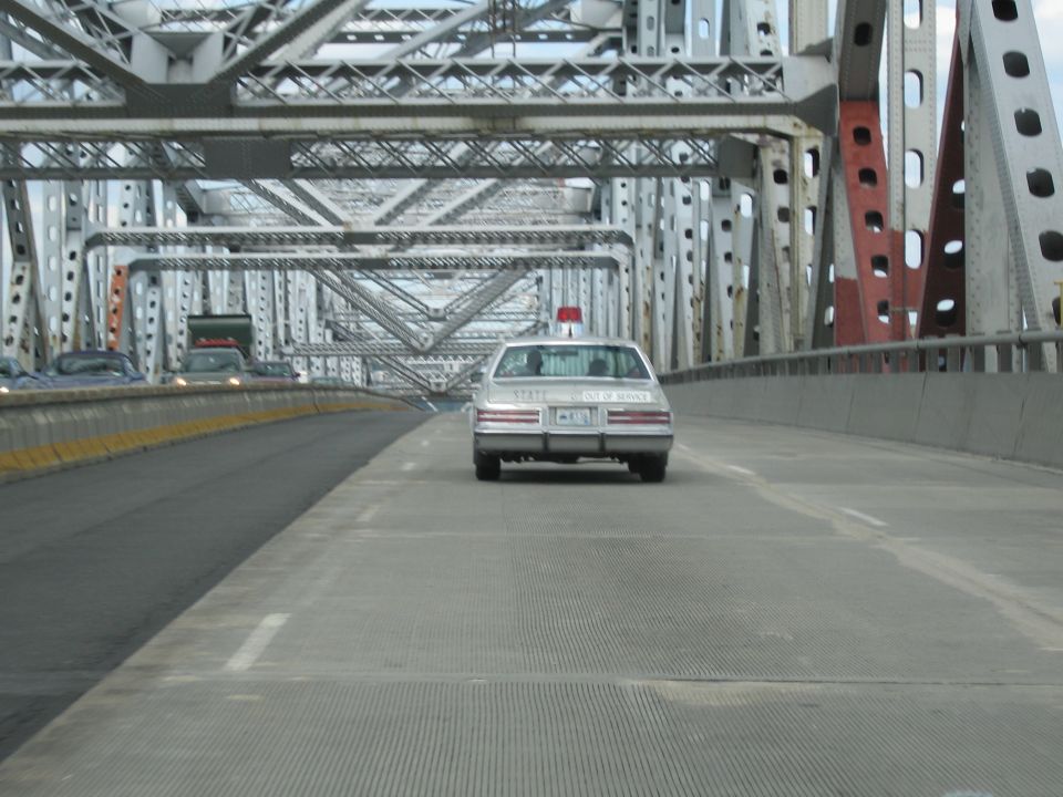 Silver state police sedan with red roof beacon driving on a steel truss bridge