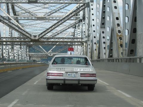 1991 Ford LTD Crown Victoria - rear of a silver State police sedan lettered STATE and OUT OF SERVICE with a red roof beacon, crossing a steel bridge.