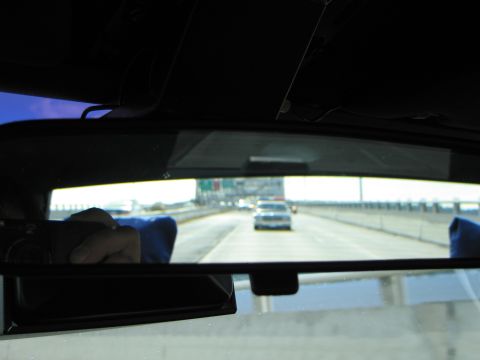 1991 Ford LTD Crown Victoria - Reflection in a car's rear-view mirror shows a police cruiser following on a highway, with a hand and blue cloth in the dim interior.