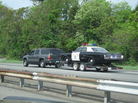 1991 Ford LTD Crown Victoria - dark gray SUV tows a black-and-white Highway Patrol sedan on a flatbed trailer along a tree-lined highway.