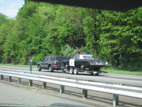 1991 Ford LTD Crown Victoria - gray SUV pulls a black-and-white police cruiser on a tandem-axle trailer along a guardrail-lined highway beside green trees.