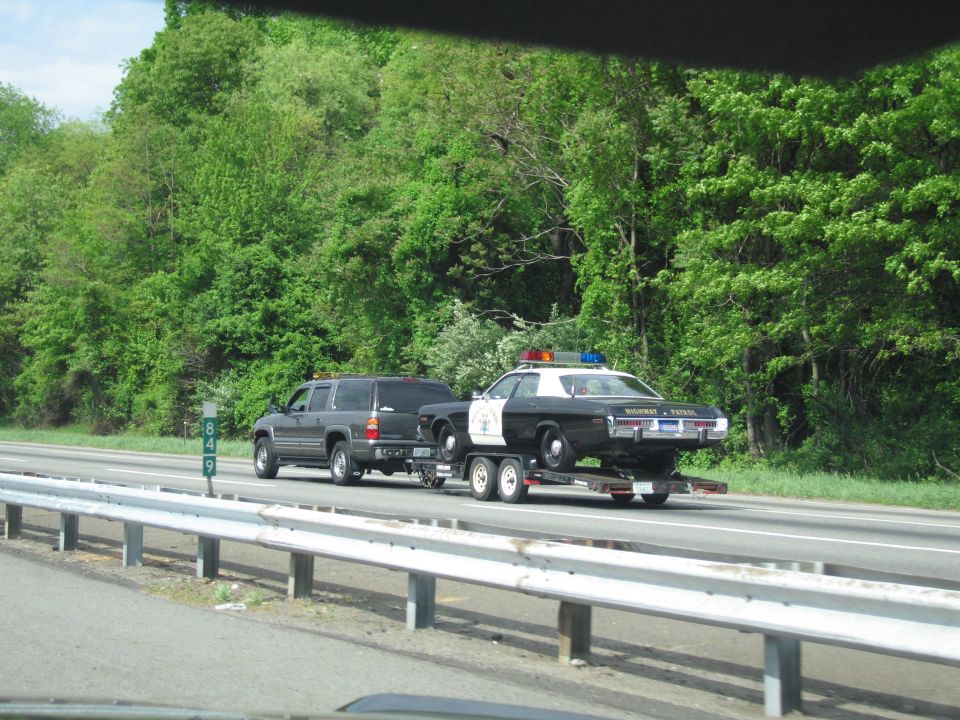 SUV hauling a black-and-white police cruiser on a trailer past green woods