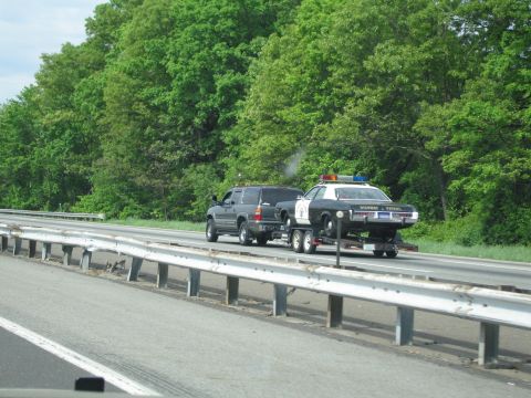 1991 Ford LTD Crown Victoria - dark SUV hauls a black-and-white Highway Patrol sedan on a flatbed trailer along a wooded interstate.