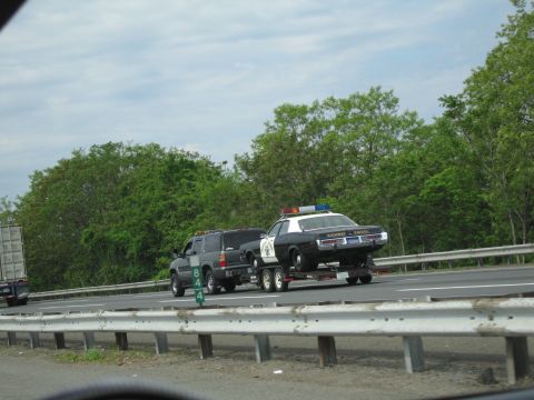 1991 Ford LTD Crown Victoria - dark SUV tows a black-and-white Highway Patrol sedan on a trailer past a green mile marker on a wooded highway.