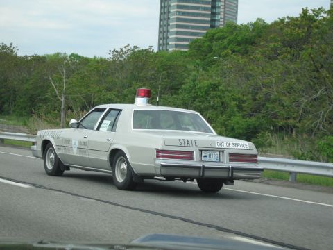 1991 Ford LTD Crown Victoria - silver State Police sedan with red roof beacon and OUT OF SERVICE lettering drives a highway with a tall building behind.