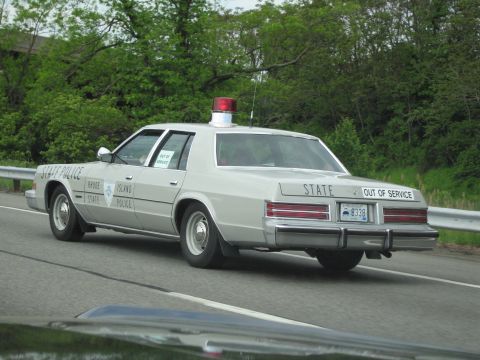 1991 Ford LTD Crown Victoria - silver sedan lettered Rhode Island State Police with a door shield, red roof beacon, and OUT OF SERVICE marking on the highway.