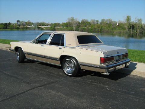 1988 Mercury Grand Marquis - A tan 1988 Mercury Grand Marquis sedan is parked on asphalt near a body of water.