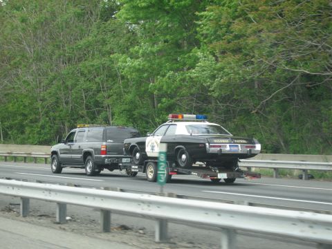1991 Ford LTD Crown Victoria - gray SUV with amber roof light tows a black-and-white Highway Patrol cruiser on a flatbed trailer along a divided highway.
