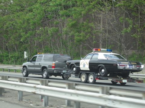 1991 Ford LTD Crown Victoria - gray SUV pulls a black-and-white Highway Patrol sedan on a tandem-axle trailer past green roadside trees.