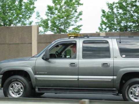 1991 Ford LTD Crown Victoria - Profile of a gray Chevrolet Suburban 2500HD with an amber roof light bar, driver visible, moving along a highway wall.