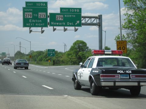 1991 Ford LTD Crown Victoria - black-and-white police cruiser with red light bar sits on a highway shoulder beneath green Exit 109 signs for Elkton and Newark Del.