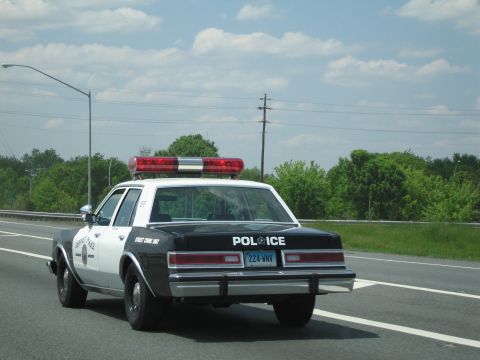 1991 Ford LTD Crown Victoria - rear of a black-and-white police sedan lettered POLICE with a red light bar, driving on a tree-lined highway.