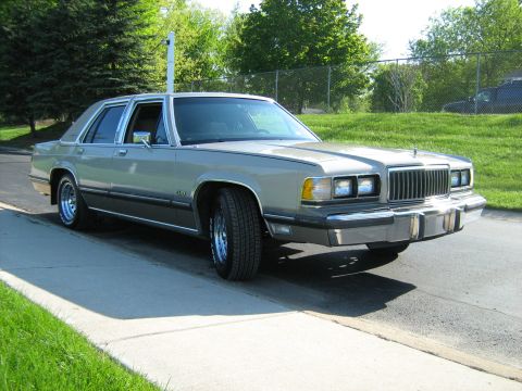1988 Mercury Grand Marquis - A silver 1988 Mercury Grand Marquis sedan is parked on a concrete surface with a grassy area and trees in the background.