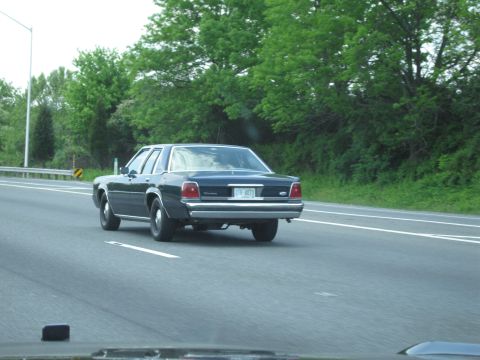 1991 Ford LTD Crown Victoria - dark blue Ford four-door sedan drives away down a highway lane beside lush green roadside trees.