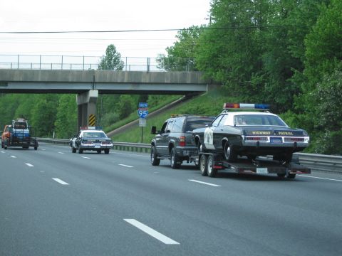 1991 Ford LTD Crown Victoria - dark SUV tows a black-and-white Highway Patrol sedan on a trailer beneath an Interstate 95 overpass, other cruisers nearby.