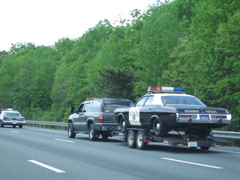 1991 Ford LTD Crown Victoria - dark SUV pulls a black-and-white Highway Patrol sedan on a trailer while a second police cruiser follows on the wooded highway.