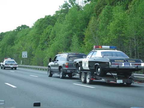 1991 Ford LTD Crown Victoria - dark SUV tows a black-and-white Highway Patrol sedan on a trailer past a Speed Limit 65 sign as another cruiser trails behind.
