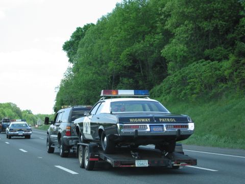 1991 Ford LTD Crown Victoria - rear of a black-and-white Highway Patrol sedan loaded on a trailer behind an SUV, with another cruiser following on the highway.