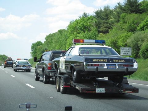 1991 Ford LTD Crown Victoria - dark SUV hauls a black-and-white Highway Patrol sedan on a trailer past an Emergency Stopping Only sign, with cruisers trailing.