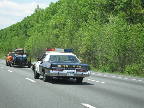 1991 Ford LTD Crown Victoria - black-and-white Highway Patrol sedan drives a wooded highway while an orange tow truck hauling another vehicle follows.
