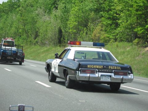 1991 Ford LTD Crown Victoria - rear of a black-and-white Dodge Highway Patrol sedan with light bar drives a wooded highway, a tow truck following.