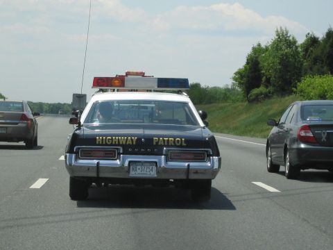 1991 Ford LTD Crown Victoria - vintage black Dodge highway patrol car with a red-and-blue roof lightbar drives down a multilane highway between other cars.