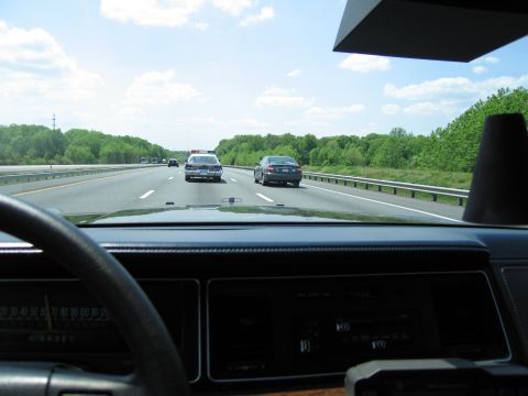 1991 Ford LTD Crown Victoria - View through a dashboard and steering wheel of a moving car, with a police vehicle and other traffic on the divided highway ahead.