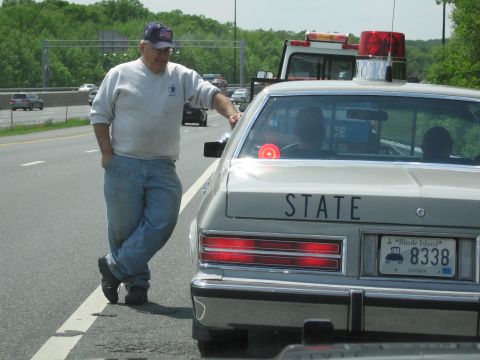 1991 Ford LTD Crown Victoria - heavyset man in a ball cap leans against a silver police sedan marked STATE, bearing a Rhode Island ANTIQUE license plate, beside a tow tru…