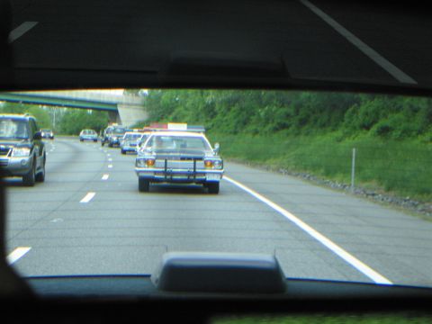 1991 Ford LTD Crown Victoria - line of classic patrol cars travels along a highway under an overpass, viewed through the rear window of a leading vehicle.