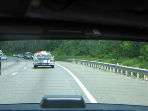 1991 Ford LTD Crown Victoria - Several patrol cars with roof lightbars travel in a row along a divided highway, seen through a car's tinted rear window.