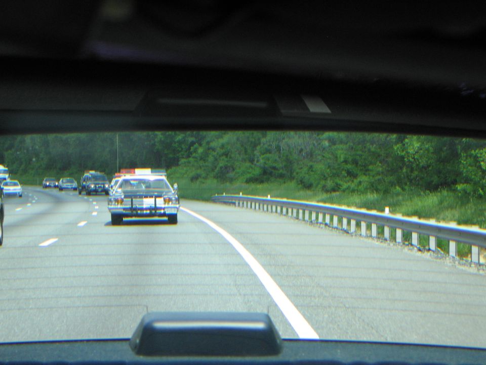 Procession of police cars on a guardrail-lined highway seen through a rear window