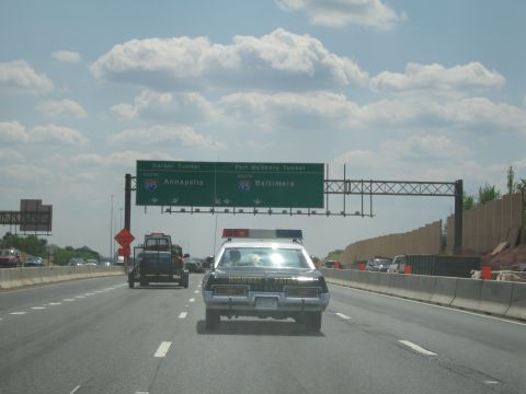 1991 Ford LTD Crown Victoria - black highway patrol sedan drives beneath green interstate signs for Annapolis and Baltimore, with a pickup towing a trailer alongside.